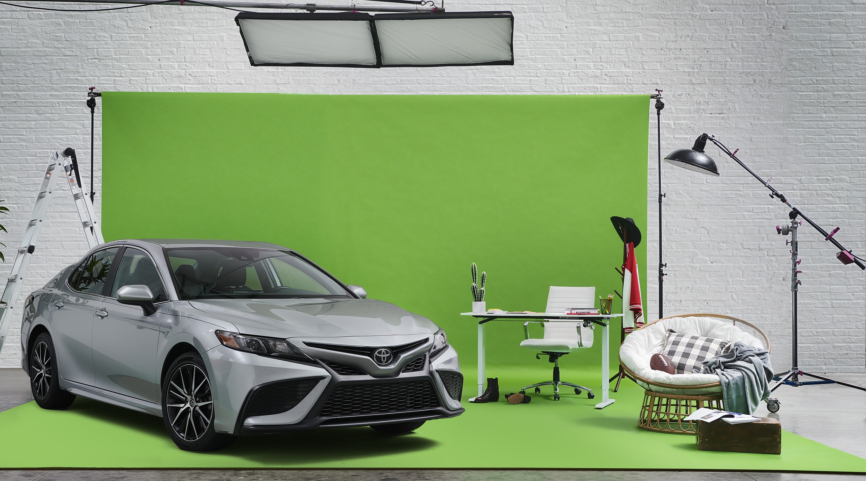 A silver Toyota Camry is parked on a greenscreen photo set with office props such as a desk and chairs.