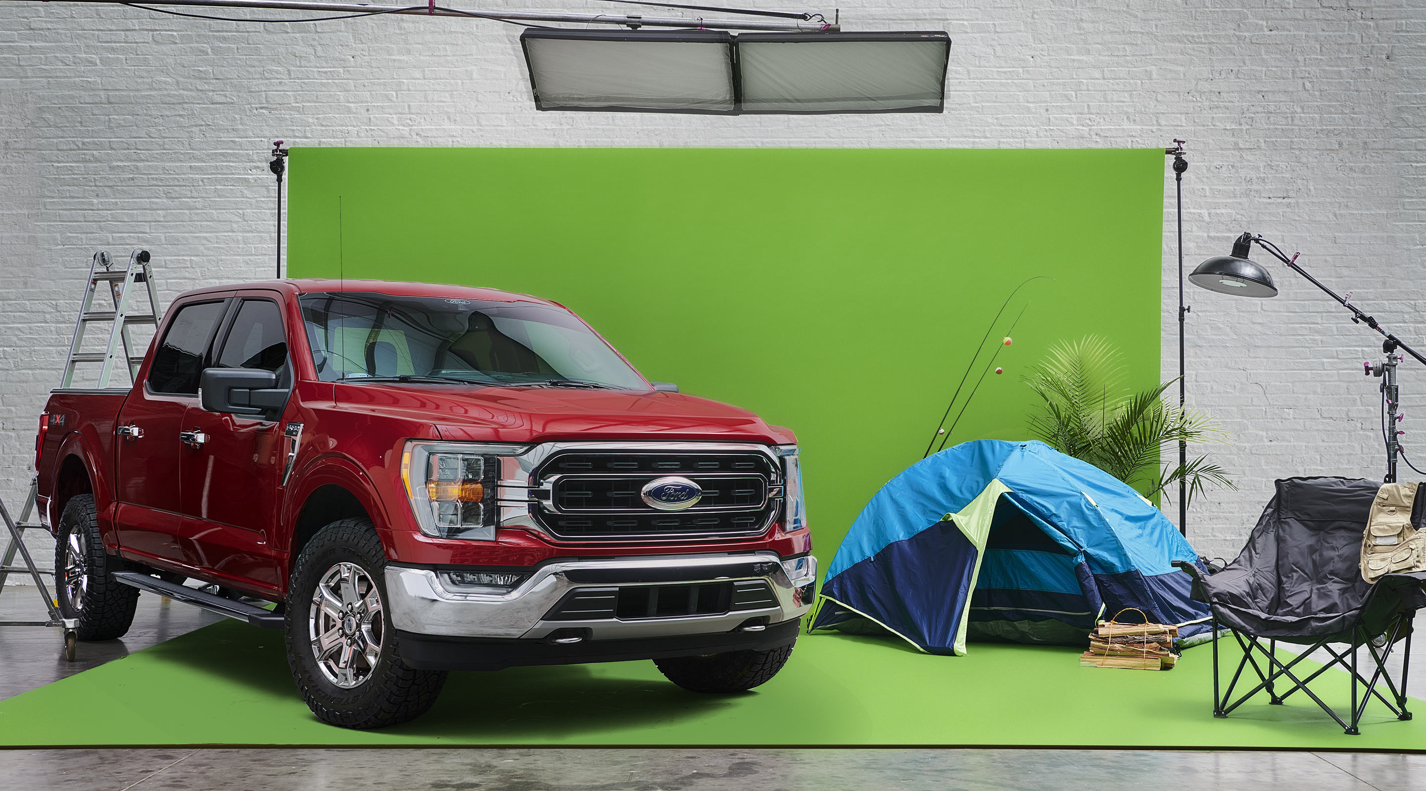 A red Ford F-150 is parked on a greenscreen photo set with outdoor props such as a tent and fishing rods.
