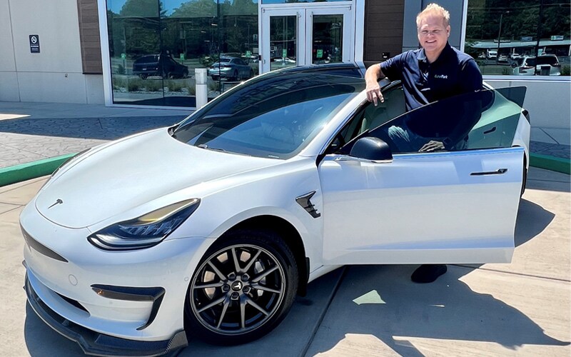 Photo shows a man named Dustin Mullinax standing in the apex of the driver’s side door of a white Tesla Model 3, parked in front of EchoPark Charlotte.