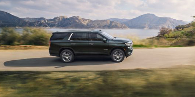 Image shows a dark gray Chevrolet Tahoe driving down a road with a lake and mountains in the background.
