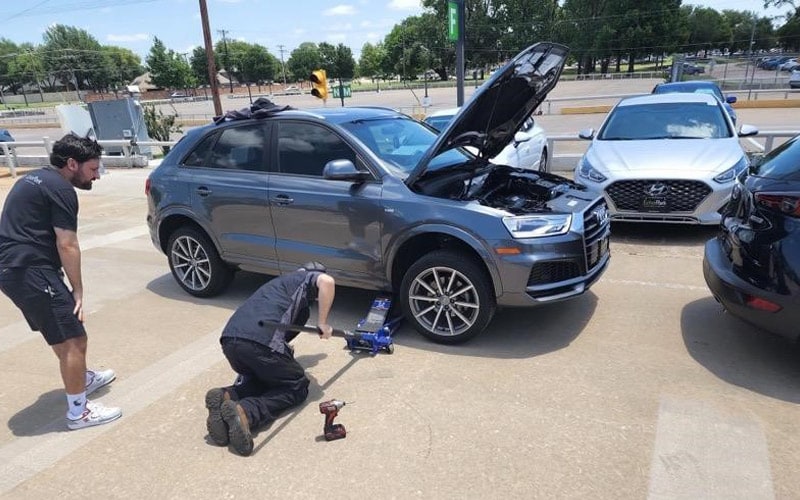 Photo shows two auto technicians using a jack to lift a light blue Audi Q3 sports utility vehicle.