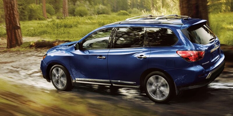 Image shows a blue Nissan Pathfinder driving along a trail through some woods.