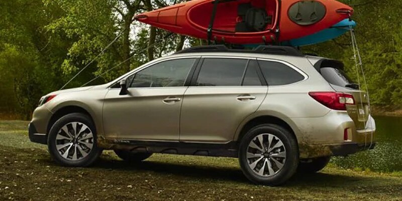 Image shows a gold Subaru Outback with a red kayak on top driving through a forested area.