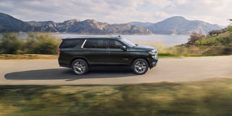 Image shows a dark gray Chevrolet Tahoe driving down a road with a lake and mountains in the background.