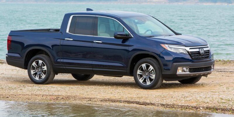Image shows dark blue Honda Ridgeline parked on sand next to water with a tree-lined shore in the background.