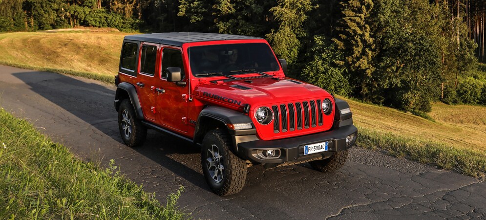 Image is of a red Jeep Wrangler with four doors and black trim, on a paved trail bordered by fields and tall trees. 