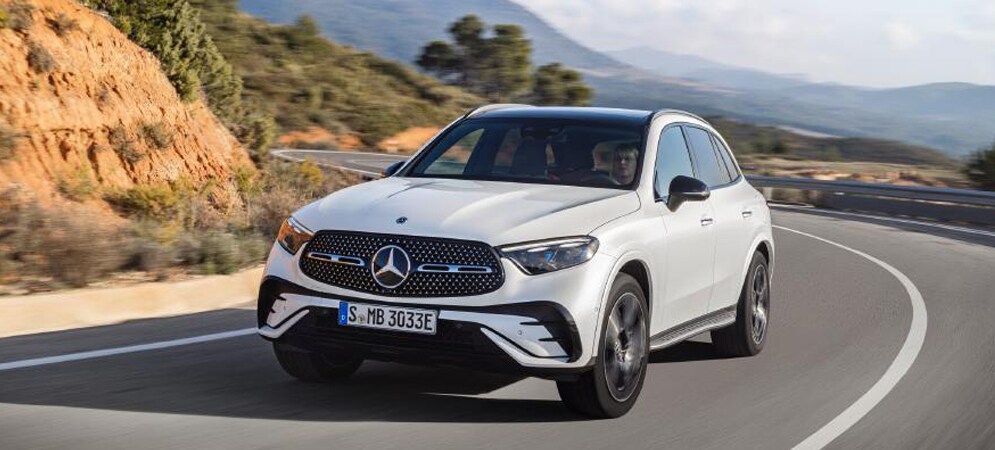 Image shows a white Mercedes-Benz GLC driving on a curved mountain roadway with a valley of hills and trees in the background.
