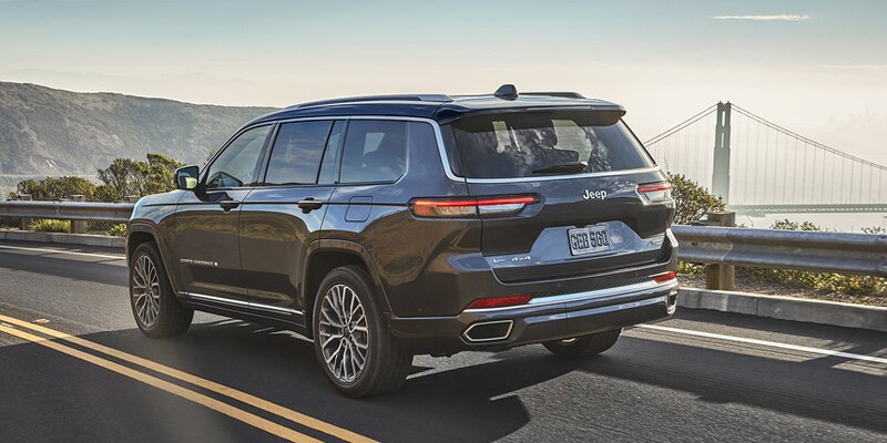 Image shows a dark gray Jeep Grand Cherokee driving on a highway, with a suspension bridge and mountains in the background.