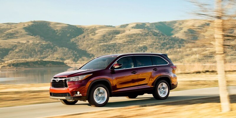 Image shows a burgundy Toyota Highlander driving down the highway, with a blurred background of mountains and hills.