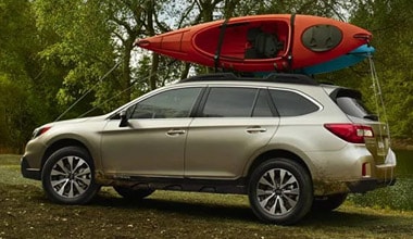 Image shows a gold Subaru Outback with a red kayak on top driving through a forested area.