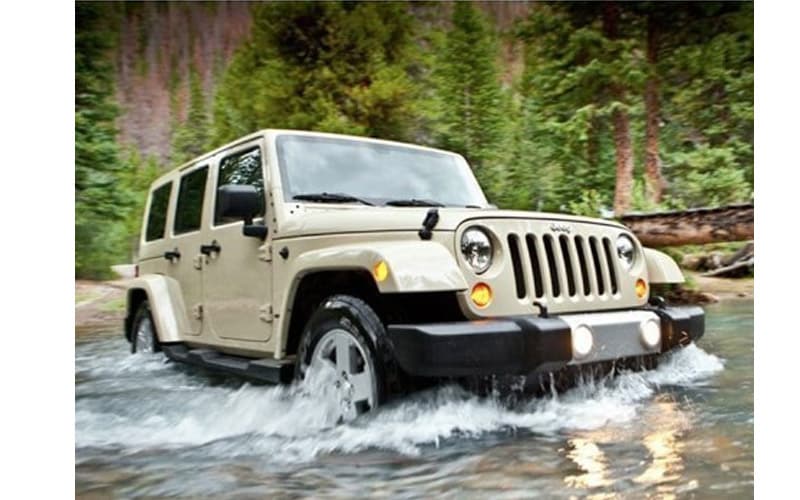 Image shows a tan Jeep Wrangler crossing a shallow river with trees in the background.