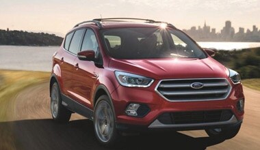 Image shows a red Ford Escape parked on a dirt road with a lake and city skyline in the background.