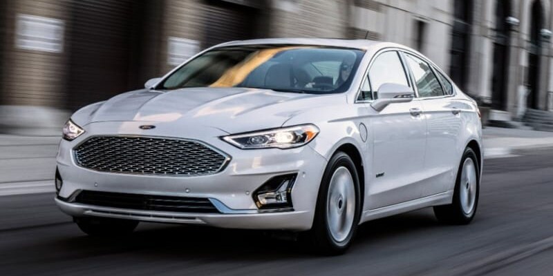 Image shows a white Ford Fusion driving down a city street.