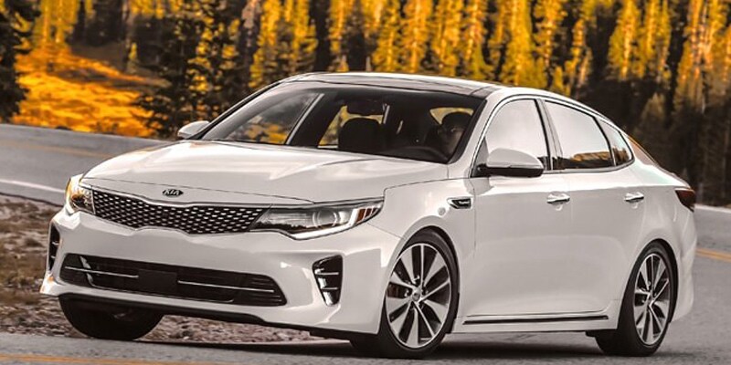 Image shows white Kia Optima driving down a highway with trees in the background.