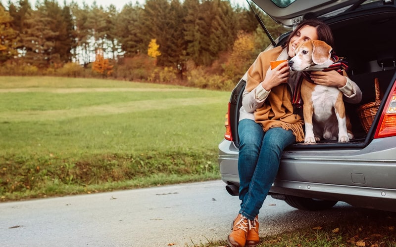 Photo shows a woman sitting with a beagle in the open liftgate of a small SUV. There is a field and trees in the background.