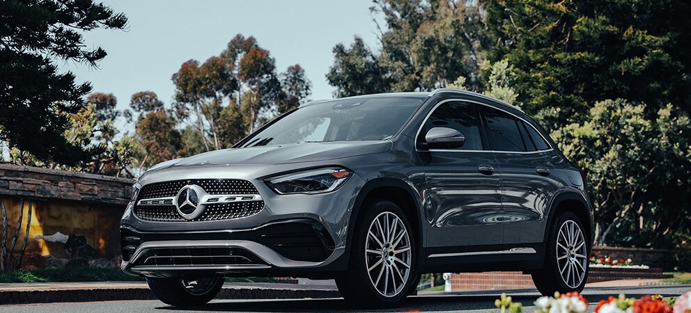 Image shows a parked gray Mercedes-Benz GLA with trees in the background and red and white flowers in the foreground.