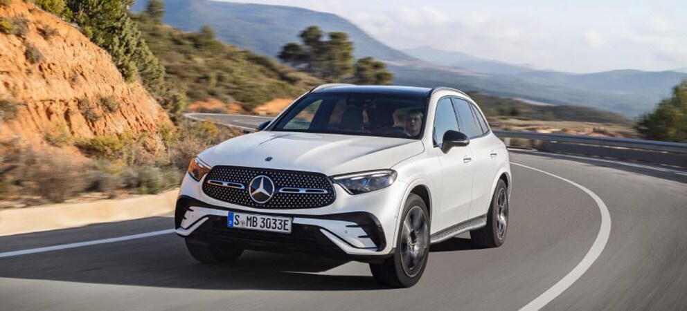 Image shows a white Mercedes-Benz GLC driving on a curved mountain roadway with a valley of hills and trees in the background.