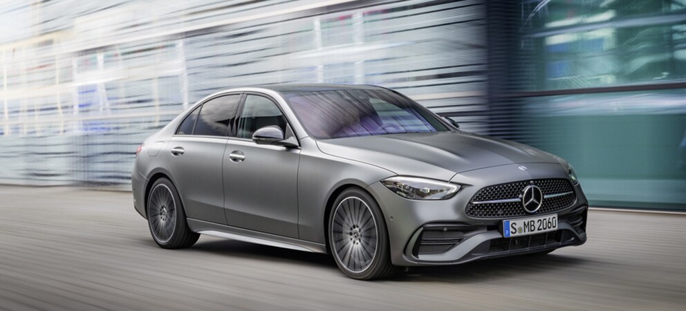 Image shows a grey Mercedes-Benz C-Class driving on a city road with blurred glass buildings in the background. 