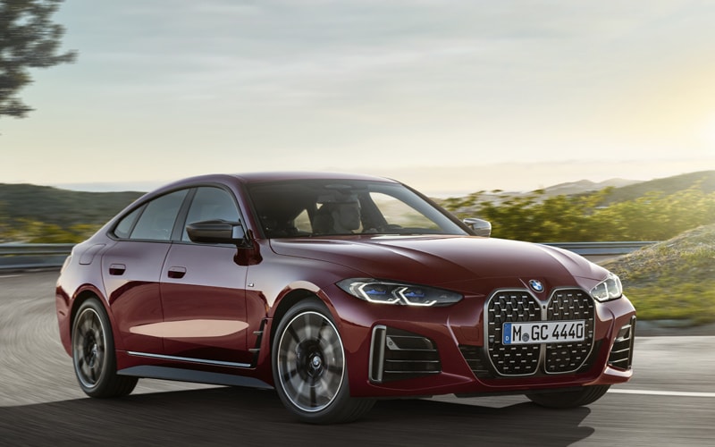 A burgundy BMW 4 Series Gran Coupe is shown rounding a curve on a paved road in hilly terrain.