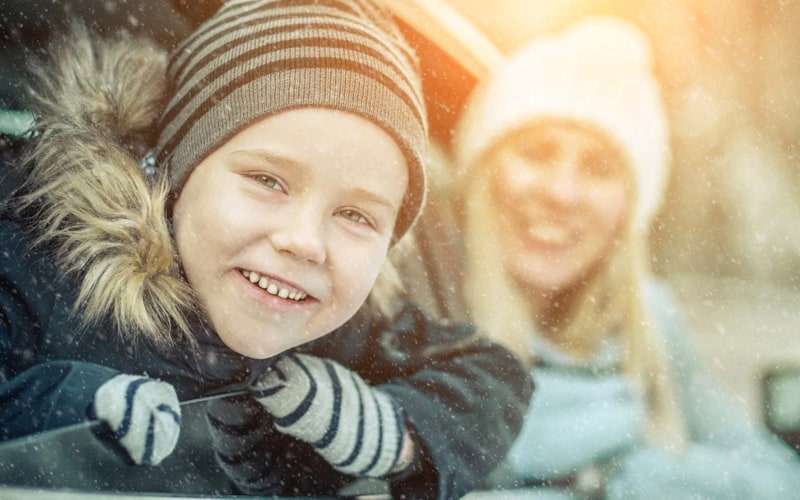 Image shows a mother and child wearing winter coats and hats leaning out a car’s windows as snow falls.