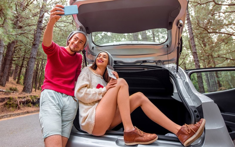 Image shows a couple in the open liftgate of a small SUV taking a selfie on a tree-lined highway.