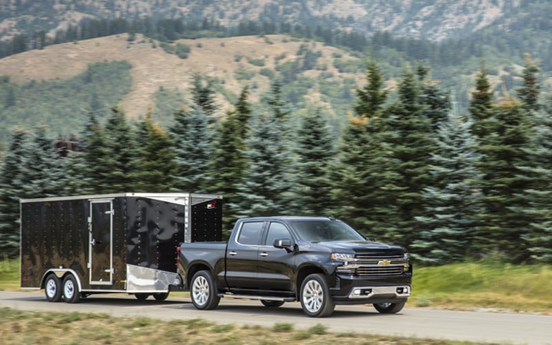 Chevrolet Silverado extended cab pulling a large tandem-axle trailer on dirt road with Evergreen trees in background. 