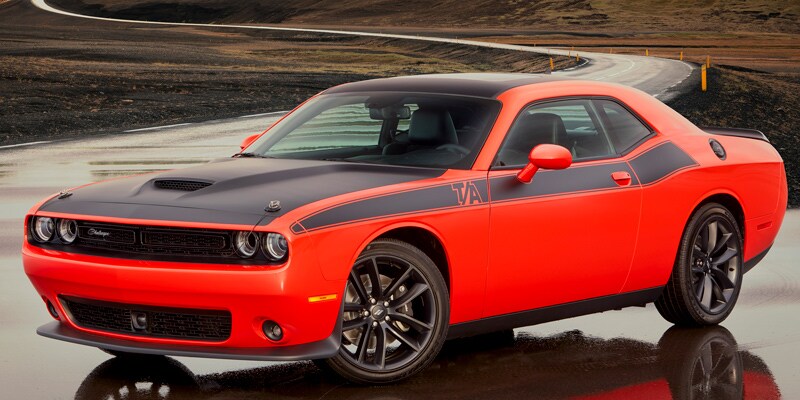 Image shows a red Dodge Challenger parked on wet, reflective asphalt with a winding road in the background. 