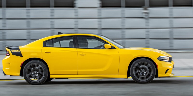 Image shows a bright yellow Dodge Charger in motion with a blurred geometric grey industrial background.