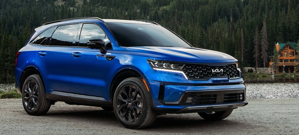 Image shows a blue Kia Sorento parks on gravel with a cabin and tress in the distance.