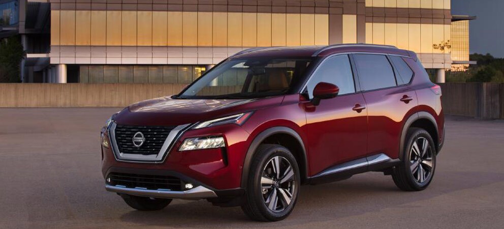 Image shows a red Nissan Rogue parked in front of a modern office building at dusk.