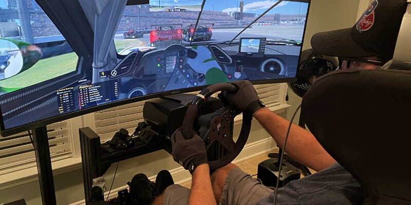 Image shows Jared DeValk, seen from the back, sitting in a gaming chair and facing a computer screen, his hands on a steering wheel. On the computer screen is the dashboard of his race car on a track.
