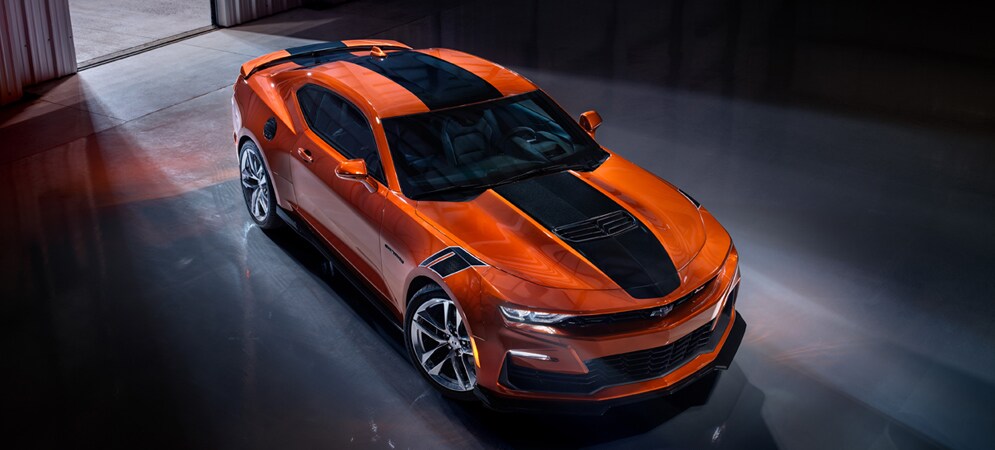Image shows orange Chevrolet Camaro with black racing stripe parked inside an industrial warehouse with dramatic lighting.