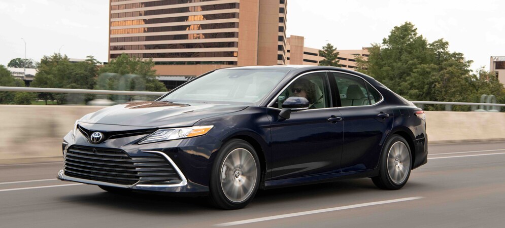 Image shows a dark blue Camry in motion, on pavement with a brown office building in the background.