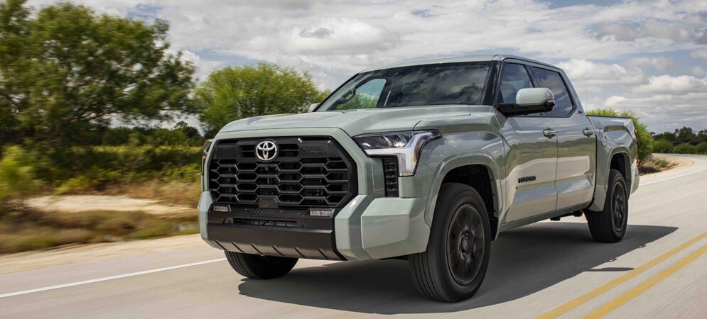 Image shows a Toyota Tundra pickup truck in a light-gray color called Lunar Rock. The truck is being driving on a divided highway, with small trees alongside the road. The sky is blue and filled with puffy, white clouds.