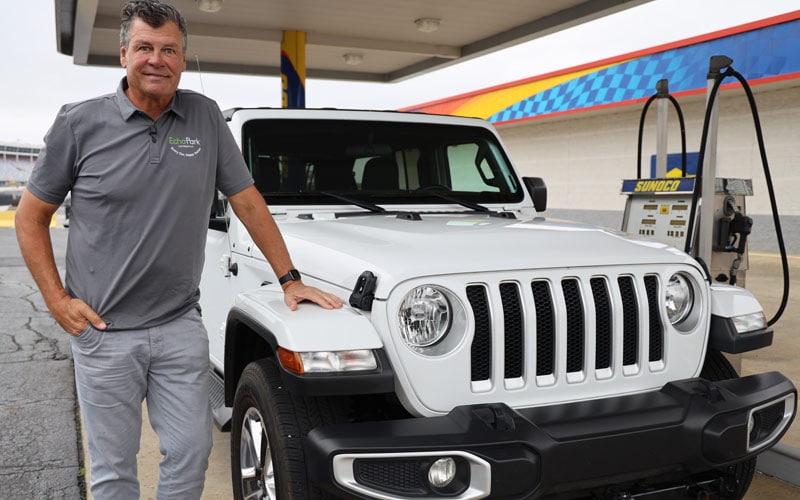 Image shows former NASCAR-driver Michael Waltrip standing by his white 2020 Jeep Wrangler Sahara. Gas pumps are in the background.
