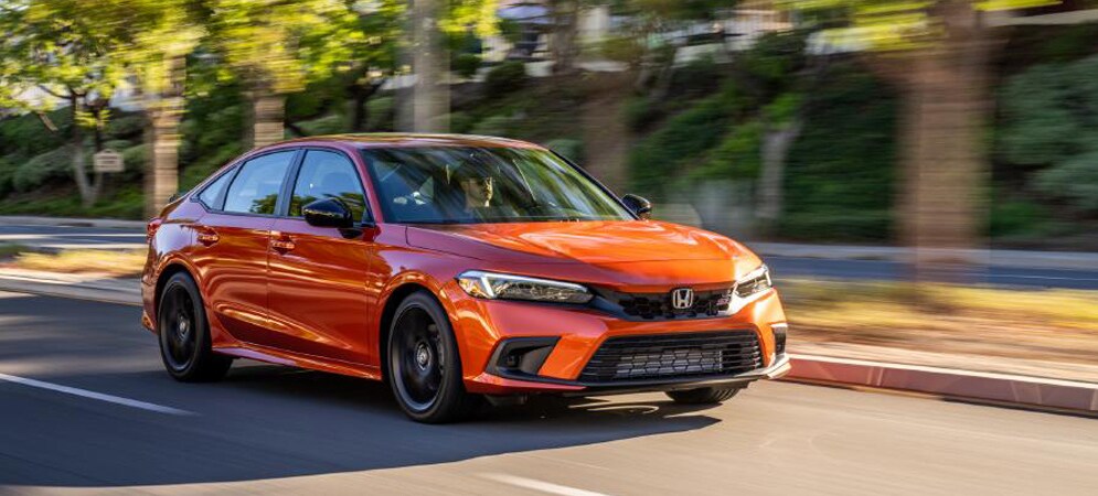 Image shows a bright orange Honda Civic driving on a road with trees in the background.