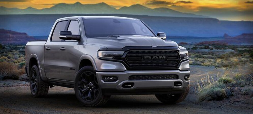 Image shows a silver Ram 1500 pickup truck in a desert landscape at sunset. There are mountains in the background.