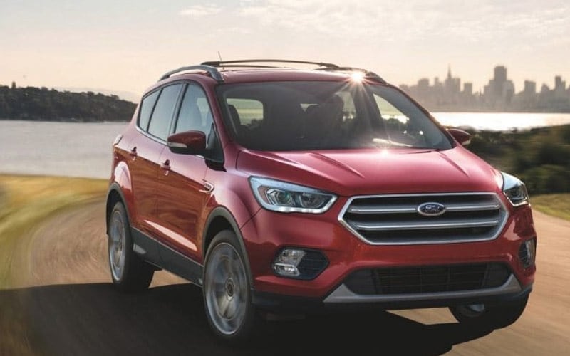 Image shows a red Ford Escape parked on a dirt road with a lake and city skyline in the background.
