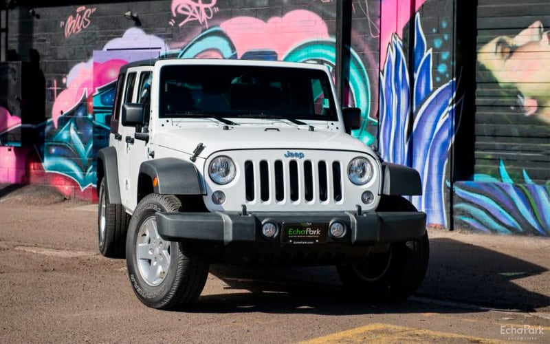 Image shows a white Jeep Wrangler parked in front of a wall adorned with a bright pink, purple and turquoise abstract mural.