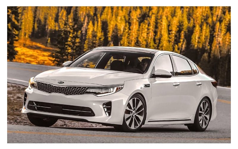 Image shows a white Kia Optima driving down a highway lined with trees that have yellow leaves.