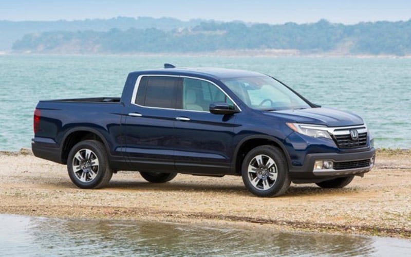 Image shows a dark blue Honda Ridgeline pickup truck parked on sand, with water and a treeline in the background.