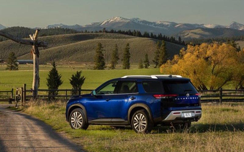 Image shows a blue Nissan Pathfinder parked on the side of a dirt road with snow-capped mountains in the background.