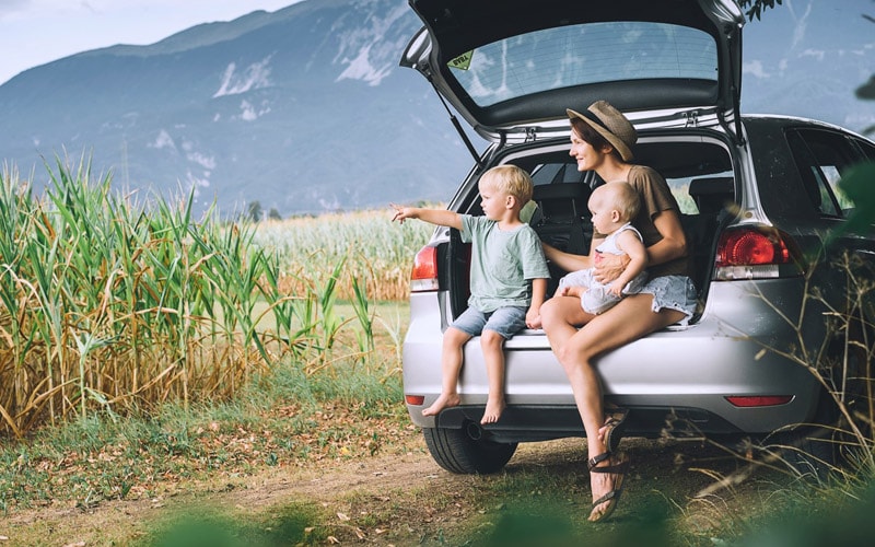 Image shows a woman sitting with a toddler and baby in the open liftgate of a SUV, with mountains in the background.