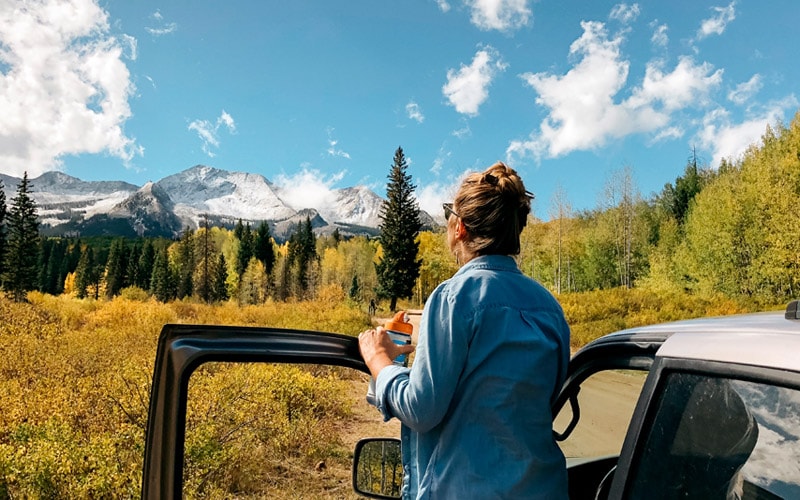 Image shows a women standing outside a parked vehicle on a dirt road looking at majestic view of mountains and trees.