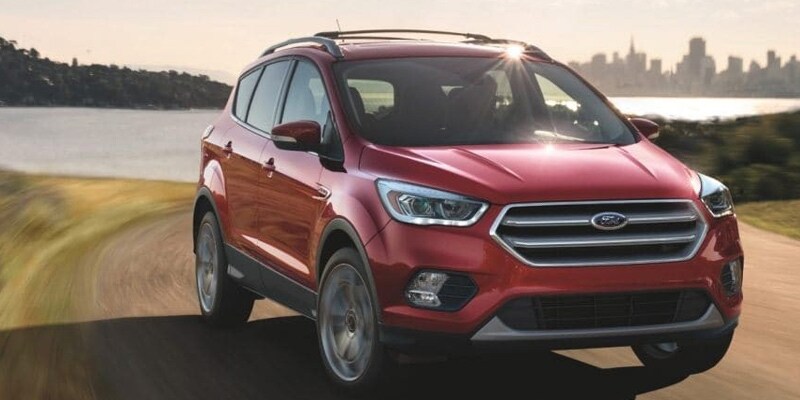 Image shows a red Ford Escape driving on a dirt road with a lake and city skyline in the background.