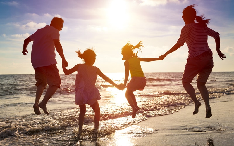 Image shows a silhouetted family of a man, woman and two girls leaping into the air at the beach.