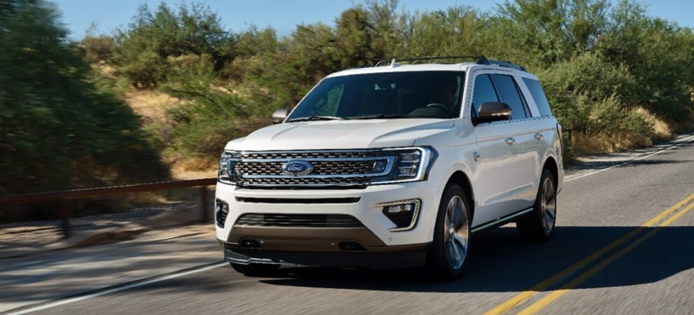 Image shows white Ford Expedition driving on a rural road, with a blurred background of hills with small trees.