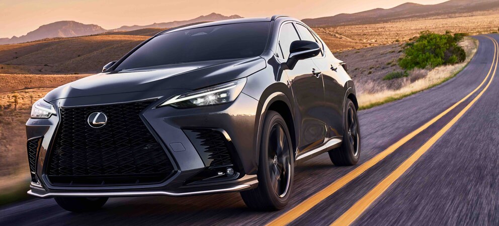 Photo shows a dark gray Lexus NX driving down a two-lane highway. It is a desert landscape with mountains in the background.