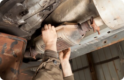 A mechanic's arm is visible as they work on a car's catalytic converter, located beneath the vehicle, in a workshop setting.