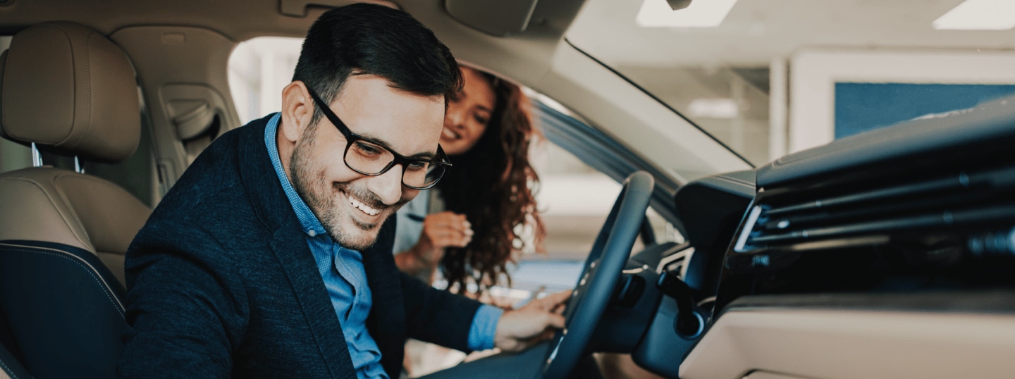 A man wearing a suit is smiling sitting in the driver's seat of a car and is getting ready for a test drive. A woman stands outside the door,  smiling and looking inside at the man.